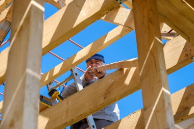 Kıdemli gri saçlı müteahhit mavi gökyüzüne karşı merdivenlerde duran ahşap bir kır evinin çerçevesini topluyor. Yaşlıların fiziksel aktiviteleri. Ahşaptan yapılmış çevre dostu evler.