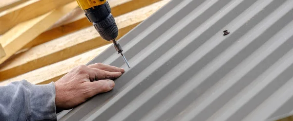 Men\'s hands in work gloves with a yellow screwdriver screw the roofing sheet to the roof of a country house. Cordless drill. The use of electrical engineering and technology.