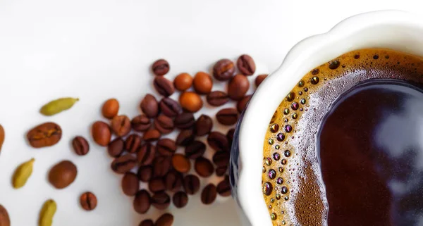 Flat lay. Banner. Black coffee with coffee foam in a white cup. In the background are coffee beans and cardamom grains on a white glossy surface. Shallow depth of field and highly blurred background.
