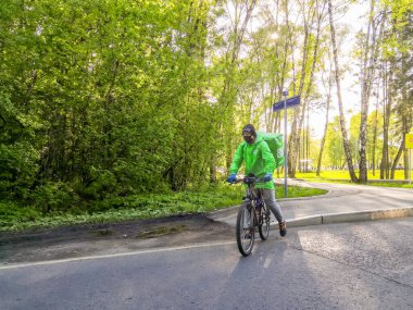 Moskova, Rusya. 15 Mayıs 2020. Güneşli bir günde, yeşil elbiseli ve siyah koruyucu maskeli bir bisikletle şehir parkında kurye. Karantina altındaki koronavirüs salgınında yiyecek teslimatı.