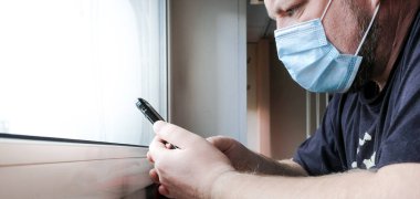 Tıbbi maskeli bir adam pencerenin önünde oturur ve endişeyle akıllı telefona bakar. Koronavirüs salgınının karantinası sırasında aile sağlığı için endişeleniyor..