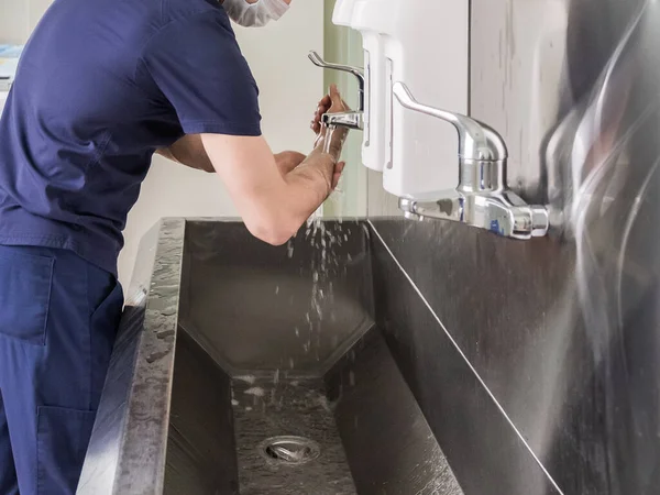 The doctor washes his hands under the tap in a stainless steel sink. Disinfection and sterilization. Removing bacteria and viruses. Necessary disinfection measures. Splashing water.
