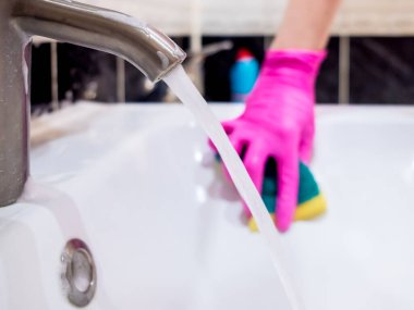 Banyodaki lavaboda musluk var. İçinden su akan yakın plan. Arka planda, pembe eldivenli bir el köpüklü deterjanla lavaboyu süngerle yıkıyor. Virüs dezenfeksiyon.