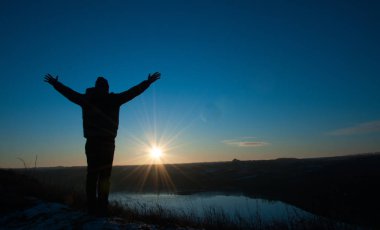 Mavi gökyüzü ve güneşli günbatımının arka planında siluetli adamlar. Elleri havada bir adam