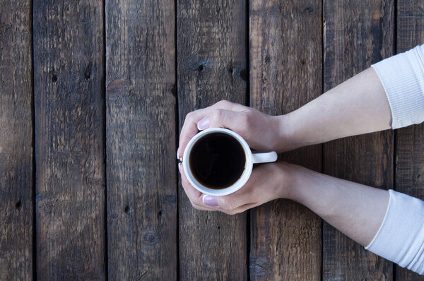 White cup with tea or coffee in the hands of a girl on a wooden background.