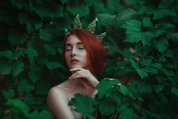 Portrait of a girl with red hair in a green leaves with a crown 