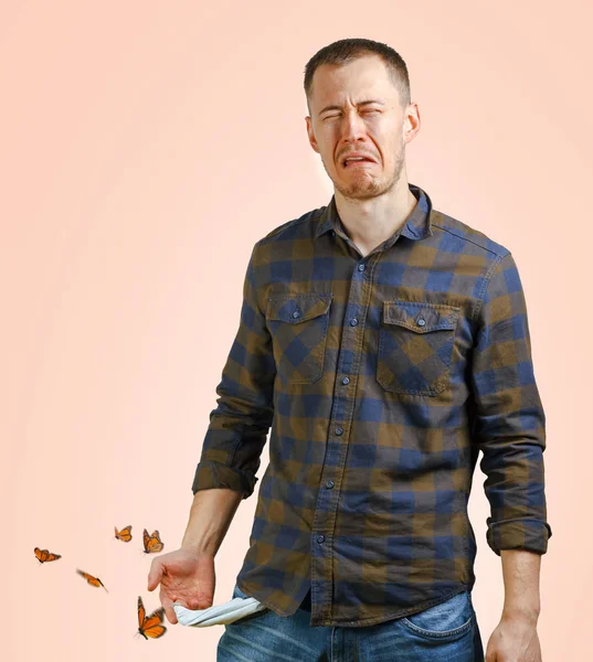 No money concept. Young man in a checkered shirt showing empty pockets butterflies fly around, sad face expression. Beige background — Stock Photo, Image