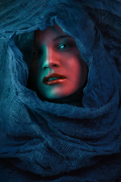Mujer cubre su cara con un paño azul, labios rojos chica bajo la capucha en luces brillantes de colores. posando en el estudio. Mujer bastante joven con bufanda con capucha — Foto de Stock