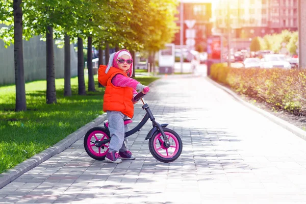 Active blond kid driving run bike the street alley of the city. Funny casual clothing vest jacket, sunglass and hood with bunny ears. Balance bike concept.