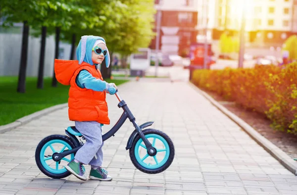 Active blond kid driving run bike the street alley of the city. Funny casual clothing vest jacket, sunglass and hood with bunny ears. Balance bike concept.