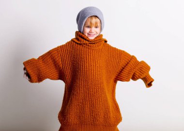 Şirin küçük bir kız, örgü şapkalı, geniş turuncu süveterli şirin bir bebek, stüdyo gri duvarında gülüyor ve oynuyor..