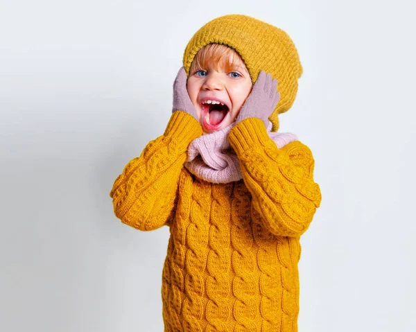 Sorpreso adorabile bambina, bambino carino in un cappello a maglia e maglione giallo, gioire rumorosamente ampiamente con la bocca spalancata sul muro grigio studio . — Foto Stock