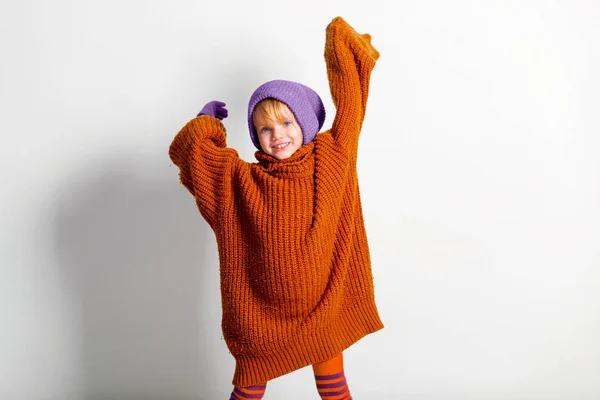 Imádnivaló kislány, aranyos kisbaba kötött sapkában és nagy narancssárga pulóverben, játszik és vicces mosolyog a stúdió szürke falán. — Stock Fotó