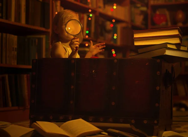 Una niña en la biblioteca de casa leyendo un libro sentado en un viejo cofre sosteniendo una lupa. Los niños y la educación muchos libros en el suelo, la noche — Foto de Stock