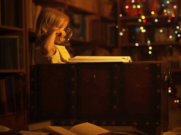 一个小女孩在家里的图书馆里看书，她坐在一个旧箱子里，手里拿着放大镜。 孩子们和教育许多书籍在地板上，深夜时分 — 图库照片
