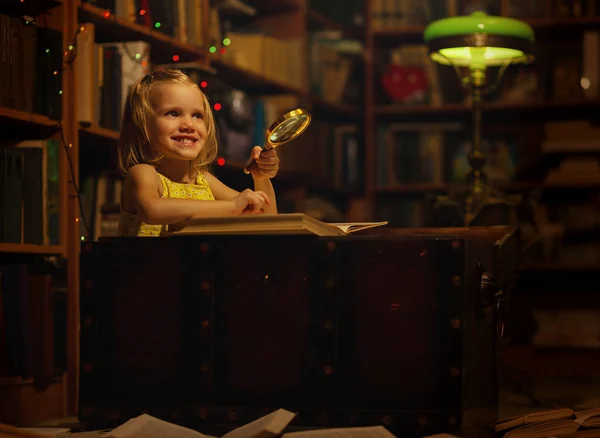 一个小女孩在家里的图书馆里看书，她坐在一个旧箱子里，手里拿着放大镜。 孩子们和教育许多书籍在地板上，深夜时分 — 图库照片