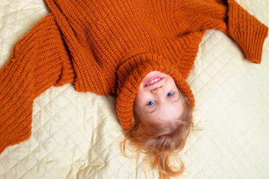 Süveter giymiş, gülümseyen ve yalan söyleyen küçük tatlı bir kızın portresi. Pek çok rahat kazağın arka planında yatıyor..