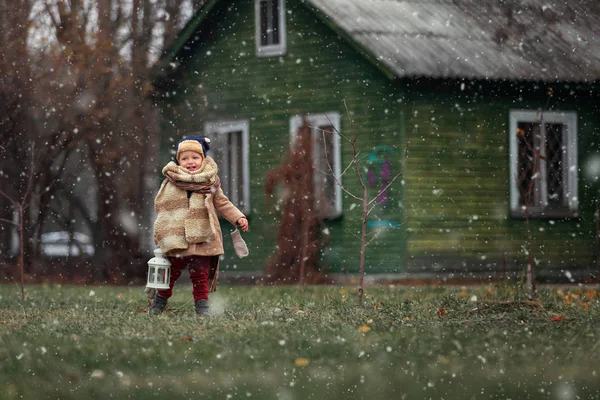 Eerste sneeuw. Kleine jongen die geniet van het platteland. Baby jongen spelen buiten met vintage handlamp, wintertijd oude houten huis op de achtergrond. — Stockfoto