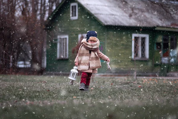 Eerste sneeuw. Kleine jongen die geniet van het platteland. Baby jongen spelen buiten met vintage handlamp, wintertijd oude houten huis op de achtergrond. — Stockfoto