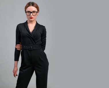 Siyah, klasik tulumlu ve gri arka planda poz veren zarif bir iş kadını. Mutlu, rahat, rahat, zarif kadın işinin tadını çıkarıyor.