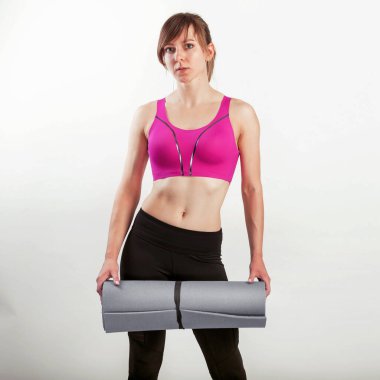 Spor kadını elinde yoga ya da spor minderi tutarken gri arka planda tek başına duruyor.