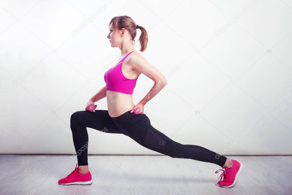 Girl workout wearing pink snickers and top, dark leggings doing lunge at gym, fitness, white wall and wooden floor, copy space.