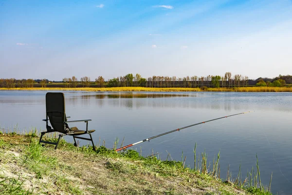 Порожній стілець і вудка на березі озера вранці. Зелена трава біля великого озера, блакитне небо, ранні години — стокове фото