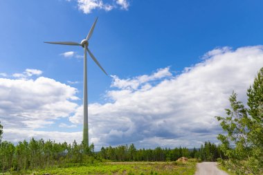 Yeşil ormanın ortasında, mavi gökyüzünün altında bulutlu tek bir rüzgar türbini. Rüzgar çiftliği, rüzgar santrali