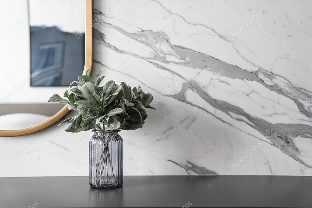 Artificial plant in glass vase compose with gold stainless mirror frame on gray spray-painted  working table with  marble wall in the background with copy space /apartment interior