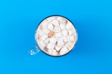 Hot cocoa in a glass mug with marshmallow on a colored blue background in the middle