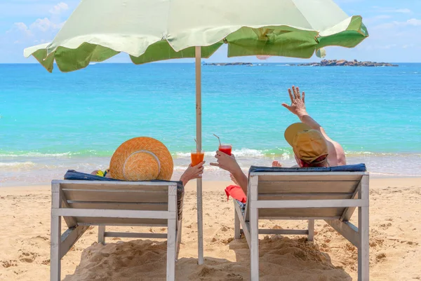 Мужчина и женщина лежат в гамаках с разноцветными коктейлями в руках. Мужчина машет рукой океану. Привет, лето. Руки с красочными коктейлями на пляже — стоковое фото