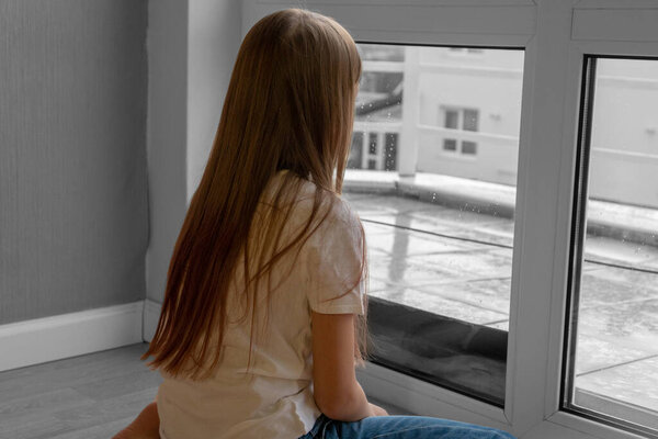 Sad teenager girl looking the rain falling through a window at home or hotel.