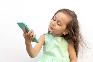 Beyaz arka planda izole edilmiş kameralı telefona gülümseyen küçük bir kız. Rüzgarda saç çıkıyor. Çekici çocuk telefonda selfie çekiyor. Modern çocuk ve moda kavramı.
