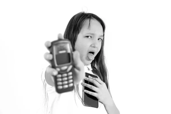 A teenager girl holds phones in her hands and looks indignantly at the camera. Tongue stuck out with displeasure. The concept of harm modern gadgets for children. Black and white photo — 스톡 사진