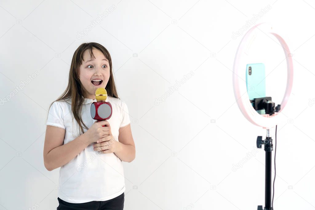A teenage girl with a microphone in her hand sings and shoots a video. Selfies. The phone is mounted on a tripod and the ring lamp shines. Emotions Girl blogger