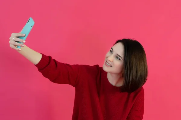 Hermosa joven con el pelo corto en un suéter rojo hace selfie en el teléfono sobre un fondo rosa brillante. Estudio foto —  Fotos de Stock