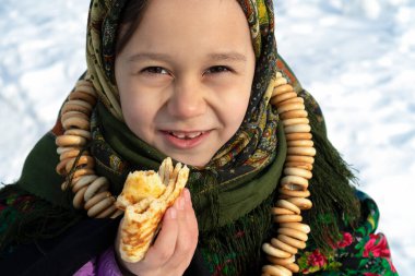 Rus halk kostümü giymiş çekici bir kız karda oturur ve krep yer. Rus bayramı kutlaması konsepti Maslenitsa