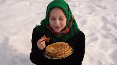 Rus halk kostümü giymiş çekici bir kız karda oturur ve krep yer. Rus bayramı kutlaması konsepti Maslenitsa