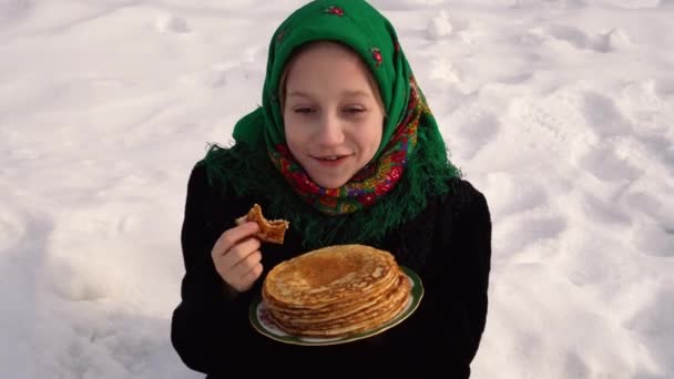 En charmig flicka i rysk folkdräkt sitter i snön och äter pannkakor. Begreppet firandet av den ryska högtiden Maslenitsa — Stockvideo