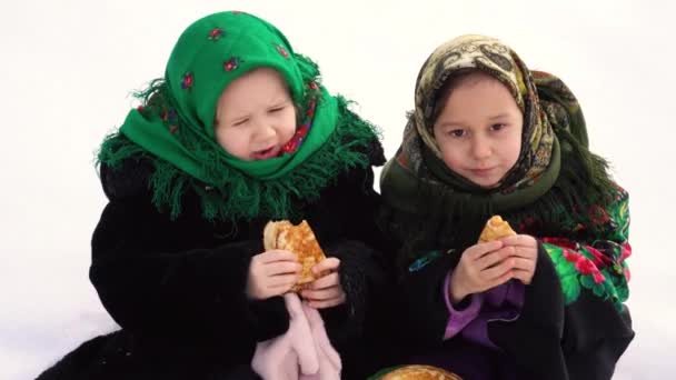 Två charmiga tjejer i en rysk folkdräkt sitter på snön och äter pannkakor. Begreppet firandet av den ryska julen shrovetide. — Stockvideo