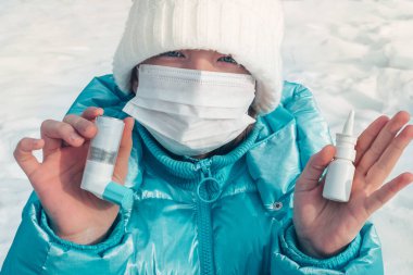 Genç kız, elinde burun ve astım spreyi için damla tutuyor. Kış parkında yürüyüş. Yüz maskesi. Grip mevsimi ve soğuk algınlığı. Alerjik çocuk. Hasta çocuklar..