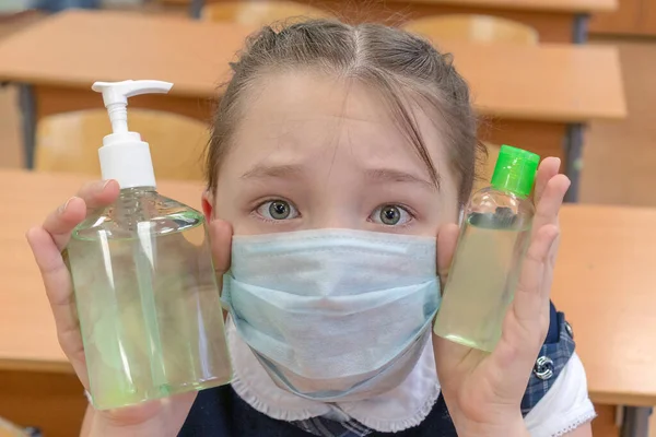 Крупним планом дівчина-школярка в медичній масці тримає в руках антисептик для дезінфекції рук у шкільному класі. Концепція захисту та гігієни дітей у зв'язку з — стокове фото