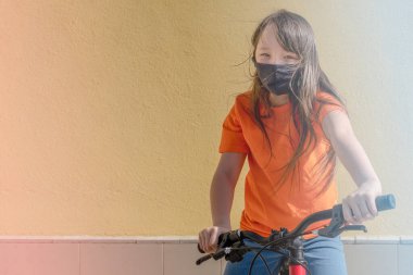 Siyah koruyucu tıbbi maskeli genç kız evin arka planında bisiklete biniyor. Coronavirus ve avluda yürüyor, kendini izole ediyor.