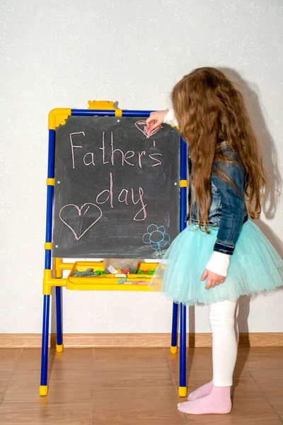 One happy girl write on the blackboard father's day and draws a heart. Concept of happy father day. Vertical photo.