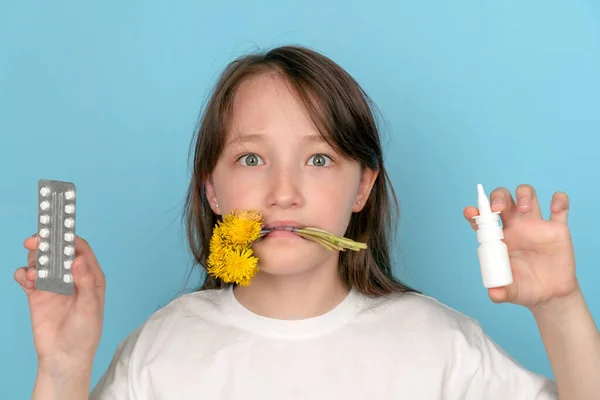 Retrato de una niña con flores en los dientes y pastillas antialérgicas y spray en las manos sobre un fondo azul. El concepto de alergia estacional al polen de las flores — Foto de Stock