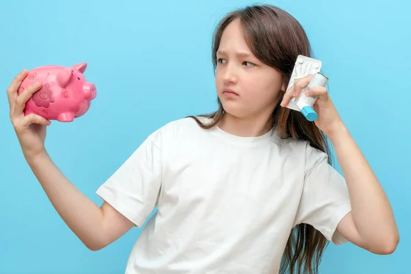 Ein 10-jähriges Mädchen hält in seinen Händen ein Sparschwein mit Geld und Medikamenten. Das Konzept der hohen Kosten für medizinische Versorgung und Medikamente — Stockfoto