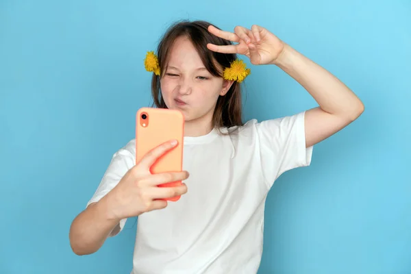 Retrato de una chica con dientes de león en el pelo y con un teléfono en las manos sobre un fondo azul. Foto del estudio. Concepto de selfie y blogger. Emoción de desprecio y el signo de Victoria —  Fotos de Stock