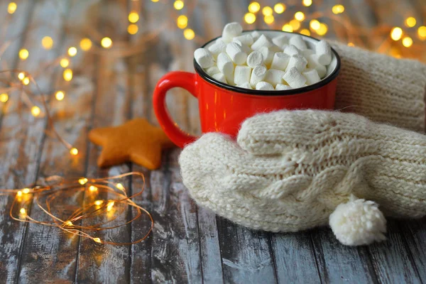 Ženská ruka v bílých rukavicích držící červený hrnek vánočního kakaa s marshmallows na dřevěném pozadí s bokeh novoroční světla. . Pijte na zimní sezónu. Comfort Food Concept — Stock fotografie