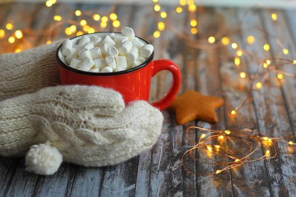 Mano de mujer en manoplas blancas sosteniendo taza roja de cacao de Navidad con malvaviscos sobre fondo de madera con luces bokeh de Año Nuevo. .. Bebe para la temporada de invierno. Concepto de Comfort Food — Foto de Stock