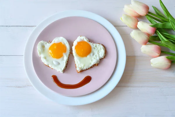 Breakfast on Valentine\'s Day - fried eggs in the shape of a heart and bouquet of pink tulips.top view